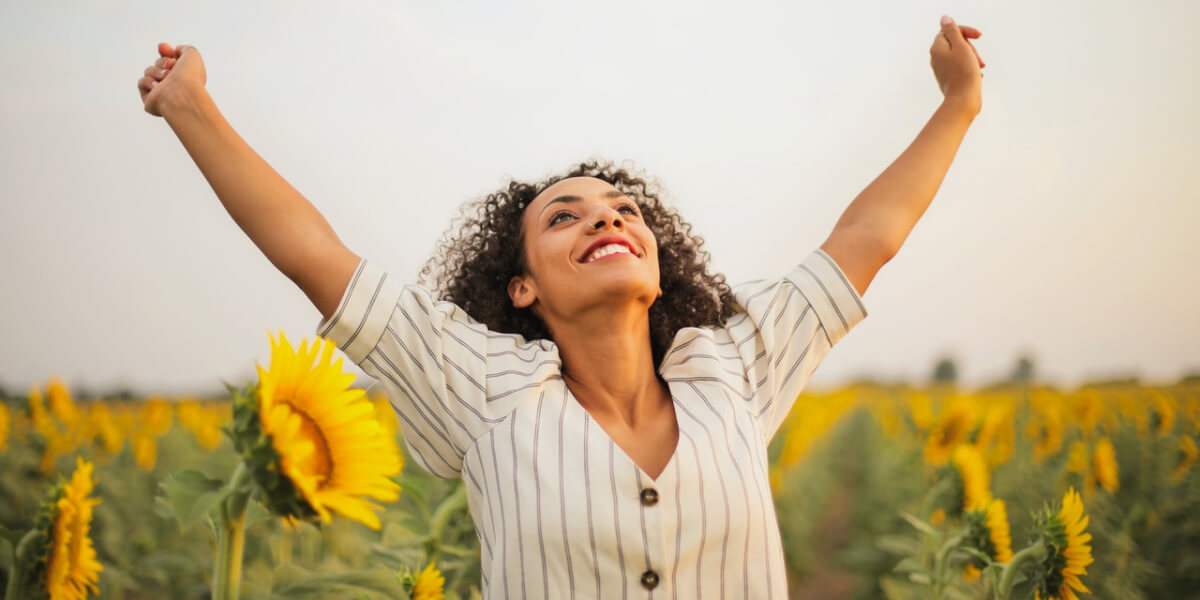 A young confident woman standing in a sunflower field with her arms raised, enjoying increased energy after eating real food.
